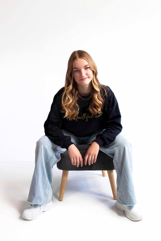 A young woman with long wavy blonde hair sits on a black stool with wooden legs, wearing a black crewneck sweatshirt with gold 'my faithful era' embroidered text, light blue wide-leg jeans, and white sneakers against a white studio background.