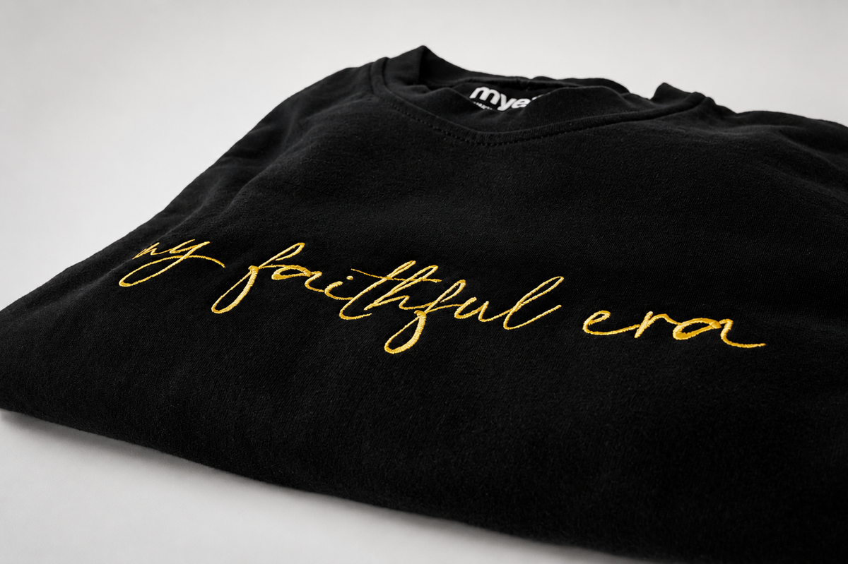 A folded black crewneck sweatshirt featuring the phrase 'my faithful era' embroidered in gold cursive script across the chest, displayed on a light gray background.