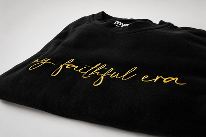 A folded black crewneck sweatshirt featuring the phrase 'my faithful era' embroidered in gold cursive script across the chest, displayed on a light gray background.