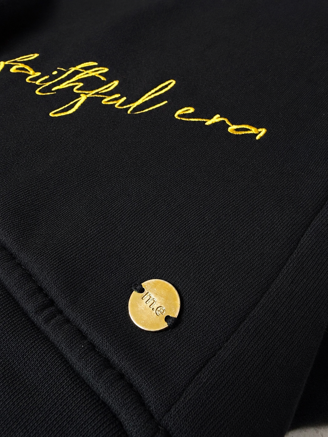 Close-up of a black crewneck sweater featuring gold embroidered script reading 'my faithful era' and a small round gold ingot with 'm.e' engraved on it. myera