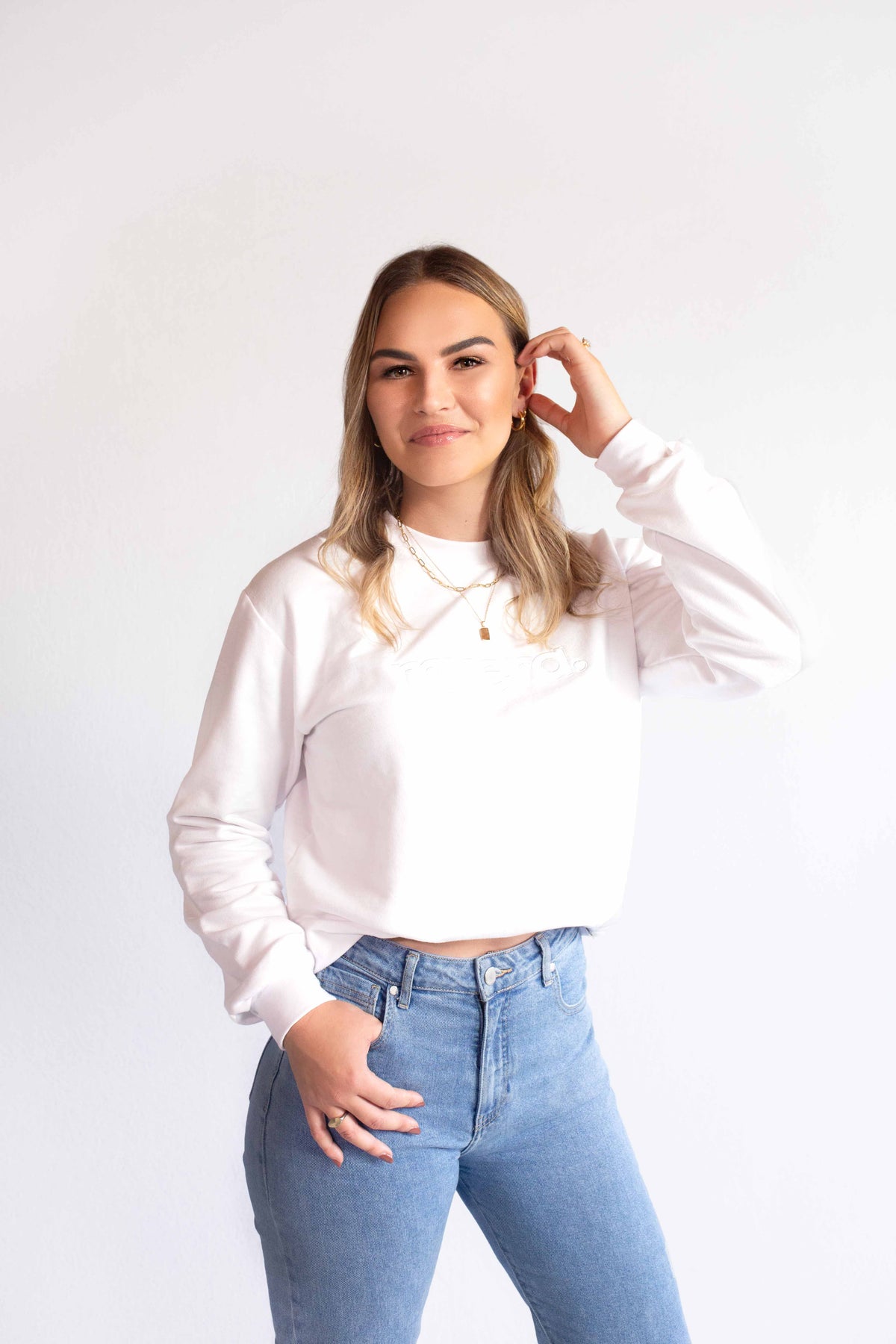 A woman modeling a white long-sleeve sweatshirt crewneck. myera embroidery on front. and light blue jeans against a plain white studio background.