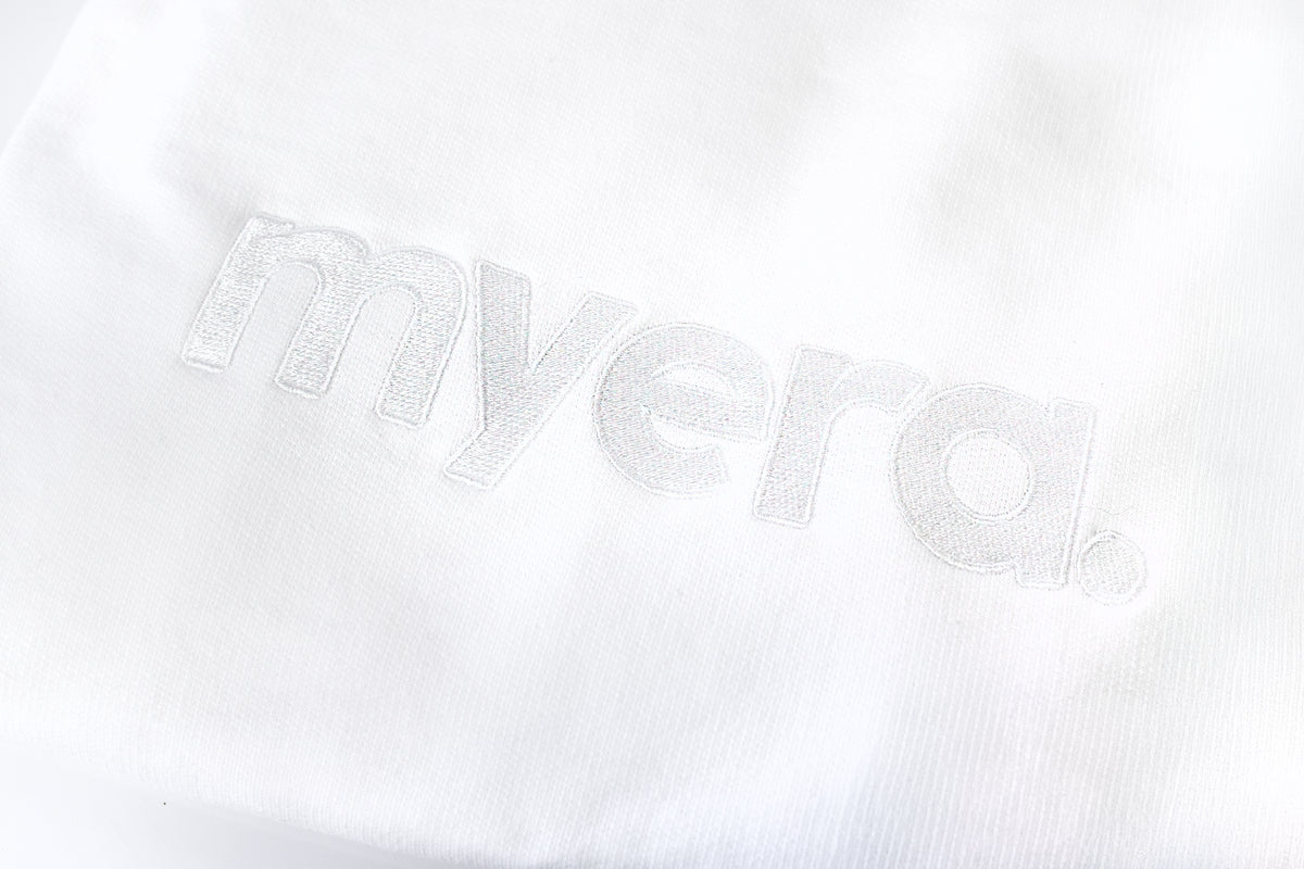 A close-up of a white textured fabric with the word 'myera.' embroidered in matching white thread. crewneck close up clothing
