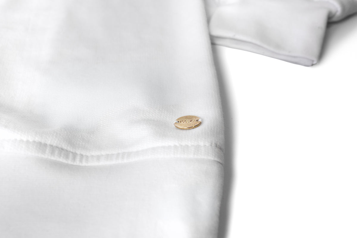 A close-up of a white cotton crewneck, a sweater, featuring a small oval gold-tone metal tag with the brand name 'myera' engraved on it. gold ingot tag