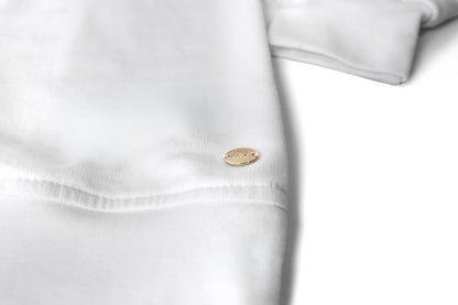 A close-up of a white cotton crewneck, a sweater, featuring a small oval gold-tone metal tag with the brand name 'myera' engraved on it. gold ingot tag