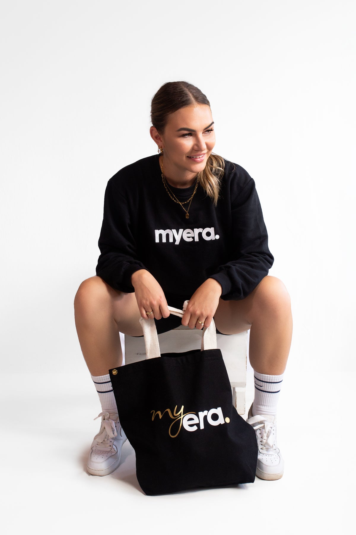 A woman sitting on a white stool wearing a black crewneck sweater with 'myera.' in white text, holding a black cotton tote bag with 'myera'.