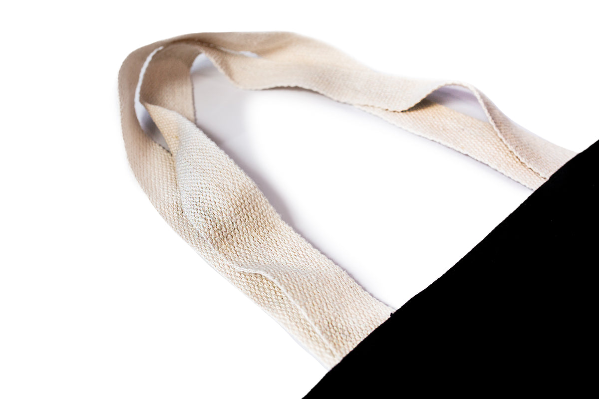 Close-up of a beige woven fabric tote bag handle, shown against a white background with a black cotton tote bag in corner. myera bag with gold ingot. printed gold and white
