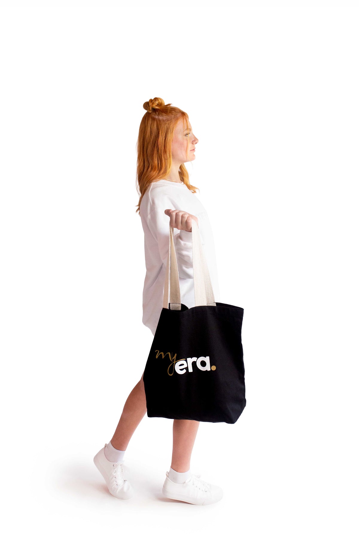 A young woman with red hair in a bun is shown from the side, wearing a white myera crewneck, sweater and white sneakers while carrying a black cotton canvas tote bag with 'my era.' printed in white and gold on the front, against a solid white background.
