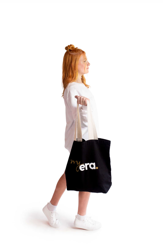 A young woman with red hair in a bun is shown from the side, wearing a white myera crewneck, sweater and white sneakers while carrying a black cotton canvas tote bag with 'my era.' printed in white and gold on the front, against a solid white background.