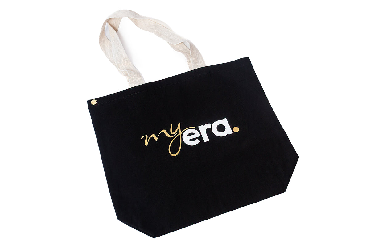 A black cotton tote bag with cream-colored fabric handles, featuring the text 'my era.' printed on the front in white and gold. myera bag