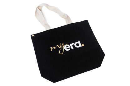 A black cotton tote bag with cream-colored fabric handles, featuring the text 'my era.' printed on the front in white and gold. myera bag