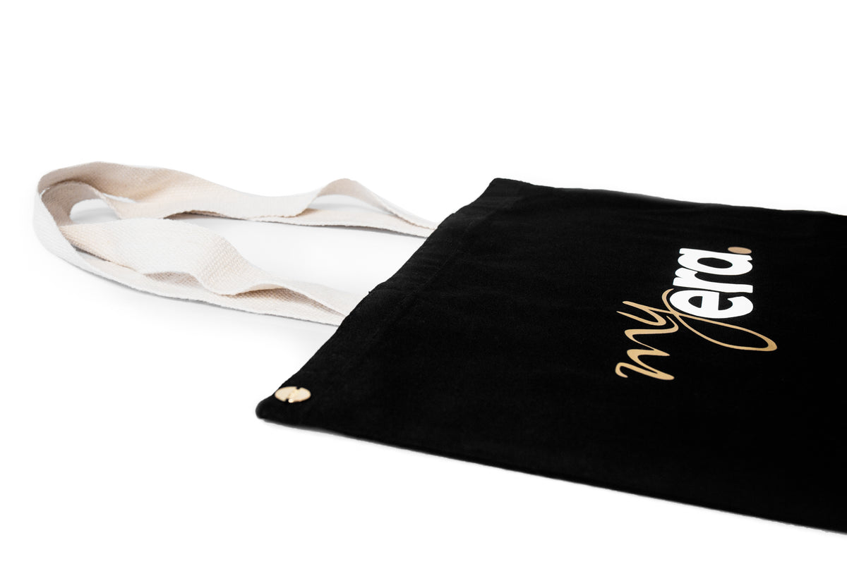 A black cotton tote bag with cream-colored shoulder straps, featuring the text 'my era.' printed in white and gold on the side, set against a plain white background. myera bag
