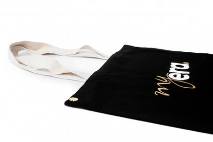 A black cotton tote bag with cream-colored shoulder straps, featuring the text 'my era.' printed in white and gold on the side, set against a plain white background. myera bag